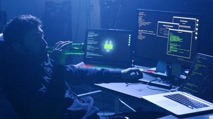 Naklejka premium Computer hacker typing typing bad data to break into computer system in the room at night. Profile of relaxed cracker drinking beer relaxing in chair at desk.