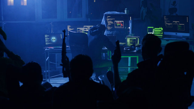 LVIV, UKRAINE - 9 JUNE, 2019: Experienced hacker typing codes on laptop to break into security system sitting at desk. Group of arrogant hackers with guns waiting in background.