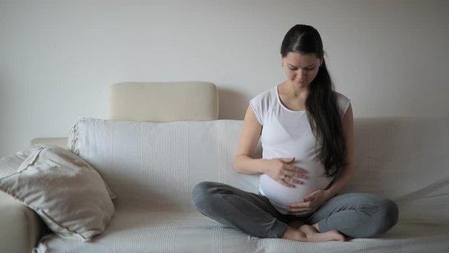 Young pregnant woman sitting on white sofa and stroking and touching belly with her hand at home