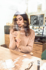 Girl holds a paper cup with coffee sitting at a wooden table in a coffee shop