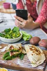 Woman take picture of vegan food with phone at her kitchen. Hand make a closeup smartphone photo of green salad and stuffed bread for blogging or social media content. Vegetarian healthy food.