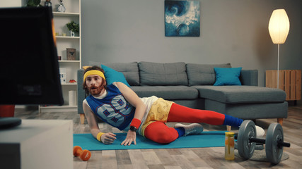 Funny-looking unsporting young man doing exercise tyrying to repeat moves watching tv in the living room at home.