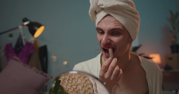 Close up of man transvestite applying pink lipstick and getting ready for party. Man transgender in loungewear sitting on sofa and looking in mirror while putting on make up.