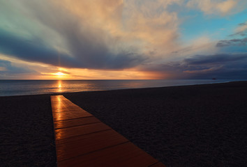 Empty beach of Calahonda in the Costa del Sol, Andalusia. Bright glowing sun rise beams reflected in wooden boardwalk, fluffy cloudy sky romantic morning or evening sunrise. Spain