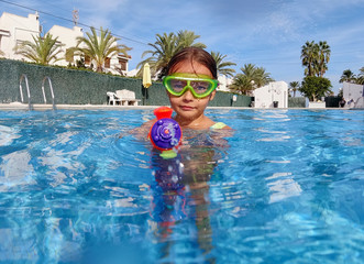 6-7 years old little adorable girl having fun in swimming pool playing aimed water gun at camera. Summer vacation relish, warm sunny weather enjoyment, playtime, carefree childhood activity concept