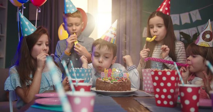 Caucasian small cute birthday boy blowing the candles on he cake while his happy friends applausing and smiling. Birthday party for children.