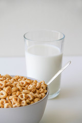 Bowl of whole grain oat cereal with a silver spoon, and a glass of milk on a white background, back view. Heart shapes cereal. 