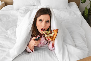 Morning of displeased young woman eating tasty pizza while watching TV in bedroom