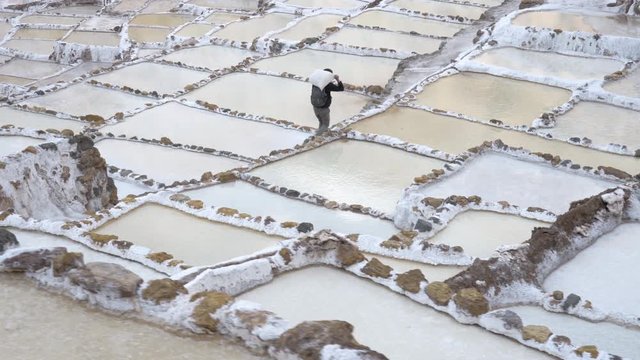 Salinas de Maras in the Sacred Valley of the Incas in the Andes mountains of Cusco region, Peru - Male worker carrying heavy bag between salt ponds