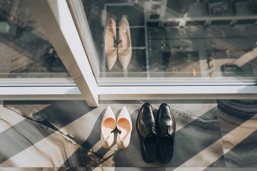 Shiny black shoes of the groom and pink shoes of bride stand near a window glass on cityscape background. Sunlight and shadow.
