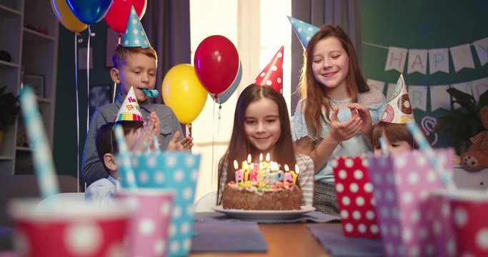 Caucasian small pretty girl in birthday conus sitting at the table among cheerful kids and blowing candles on the cake at her birthday party for friends.