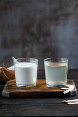 A glass with coconut milk, a glass with coconut water on a wooden board, slices of coconut nearby.