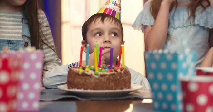 Close up of the cute little Caucasian boy in a birthday conus sitting in front of the cake with candles and eating piece with cream.