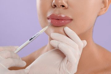 Young woman getting lips injection on lilac background, closeup