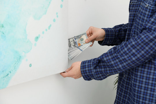 Man hiding dollar banknotes behind painting indoors, closeup. Money savings