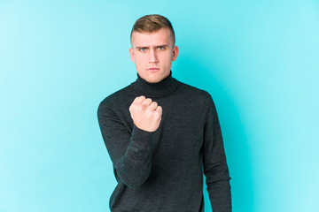 Young caucasian man on a blue background showing fist to camera, aggressive facial expression.
