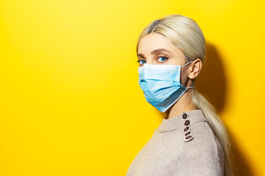 Studio Profile Portrait Of Young Blonde Girl Wearing Medical Flu Mask And Sweater On Yellow Background With Copy Space.