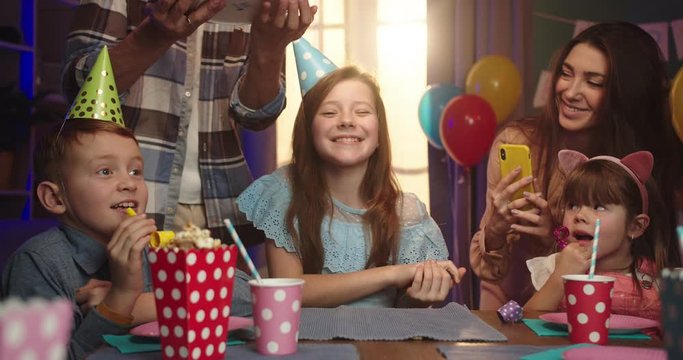 Caucasian pretty birthday girl in conus sitting at the table with friends while father or older brother coming and bringing birthday cake.