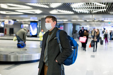 Young European man in gray coat, protective disposable medical mask in airport. Afraid of dangerous N-CoV 2019 influenza coronavirus mutated and spreading in China. Blue backpack, suitcase on wheels