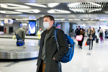Young European man in gray coat, protective disposable medical mask in airport. Afraid of dangerous...
