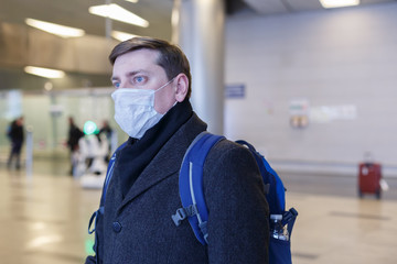 Young European man in gray coat, protective disposable medical mask in airport. Afraid of dangerous N-CoV 2019 influenza coronavirus mutated and spreading in China. Blue backpack, suitcase on wheels