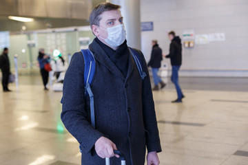 Young European man in gray coat, protective disposable medical mask in airport. Afraid of dangerous N-CoV 2019 influenza coronavirus mutated and spreading in China. Blue backpack, suitcase on wheels