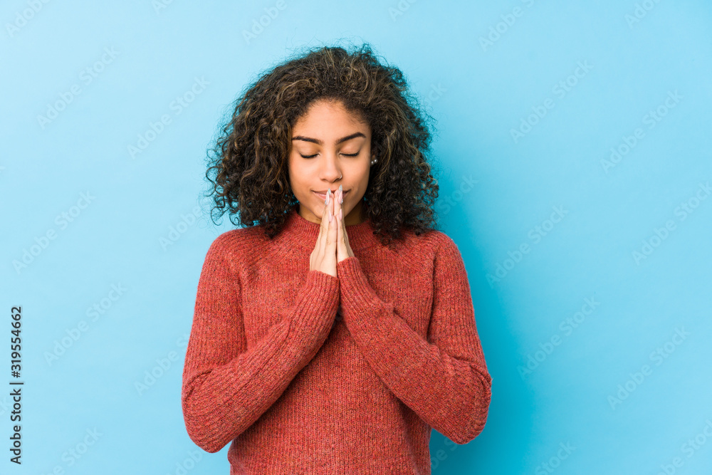 Wall mural young african american curly hair woman holding hands in pray near mouth, feels confident.