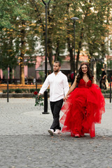 Beautiful romantic couple. Attractive young woman in red dress and crown with handsome man in white shirt with red rose walking on the street.Happy Saint Valentine's Day. Pregnant and wedding concept.