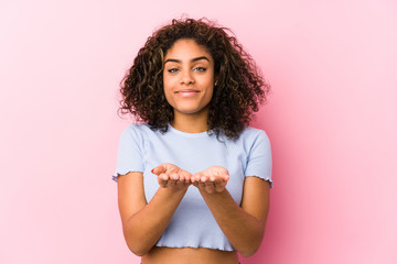 Young african american woman against a pink background holding something with palms, offering to...