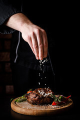 The concept of cooking meat. The chef cook salt on the cooked steak on a black background, a place under the logo for the restaurant menu. food background image, copy space text