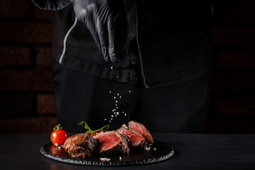 The concept of cooking meat. The chef cook salt on the cooked steak on a black background, a place...