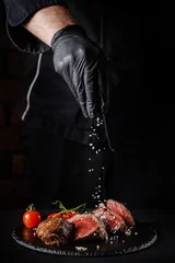 Poster The concept of cooking meat. The chef cook salt on the cooked steak on a black background, a place under the logo for the restaurant menu. food background image, copy space text © zukamilov