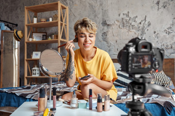 Make-up Blogger. Young woman sitting at stylish apartment recording video on camera applying...