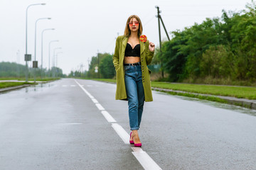 young european girl walks in rainy weather down the street with puddles of green cloak and eats...