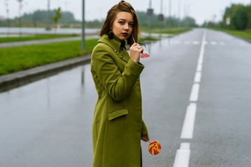 young european girl walks in rainy weather down the street with puddles of green cloak and eats...