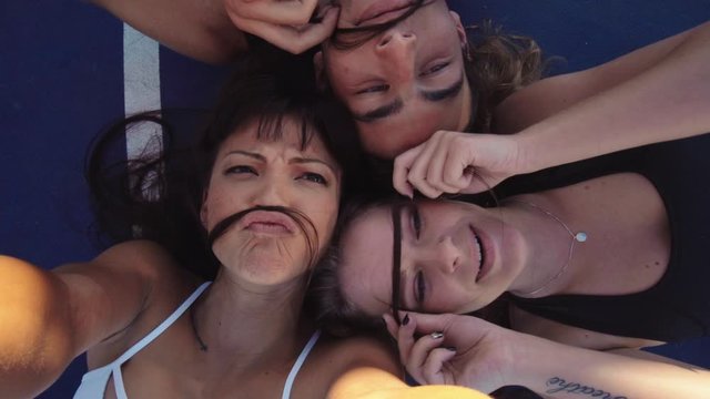 Top view of young friends lying together making fake mustache with hair and making a selfie video. Group of friends enjoying themselves outdoors.