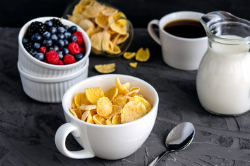 Healthy breakfast with cornflakes in a white cup, berries, milk and coffee on a dark gray background.