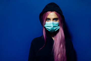Studio portrait of young girl with pink hair and blue eyes, wearing medical flu mask and hooded...