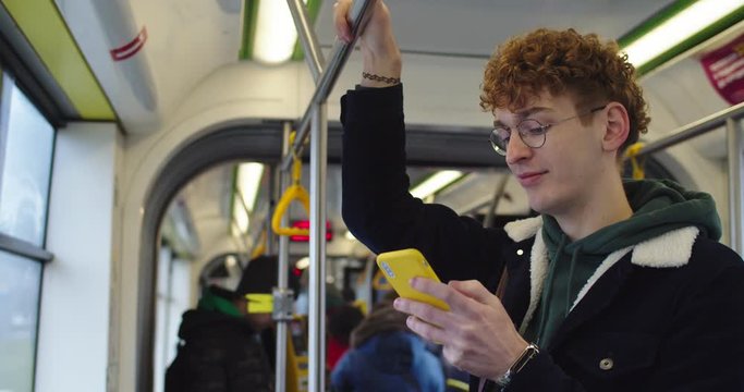 Caucasian young guy in glasses and with red hair texting a messsage while chatting on the smartphone in the tram or bus and smiling.