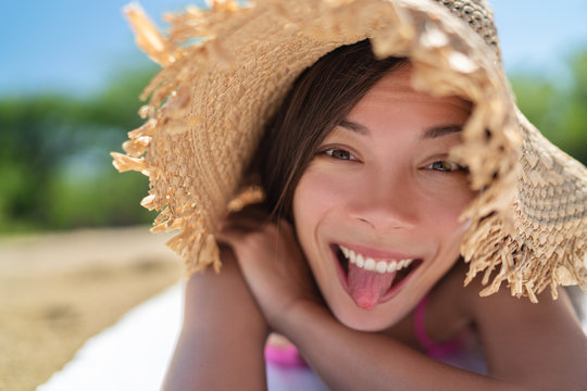 Happy funny face Asian young woman doing goofy facial expression sticking tongue out on summer vacation relax holiday sun tanning on beach having fun.