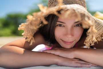 Asian woman lying down tanning wearing straw hat as sun protection on beach holiday. Summer travel beautiful young biracial girl relaxing smiling. - Powered by Adobe