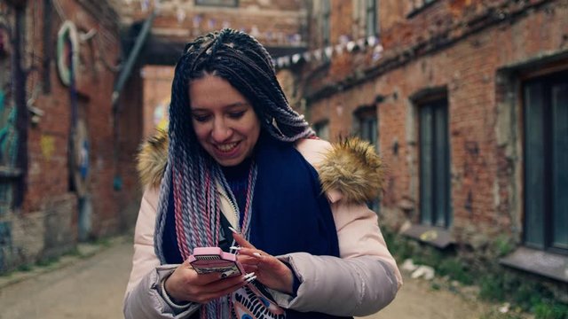 Bright young woman with dreadlocks smiles with phone. Stock footage. Young woman in colorful modern image sits on her phone and laughs on background of old streets