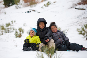 A latino grandmother hugging her grandson and granddaughter while sitting in a snow covered mountain, wearing winter clothing.
