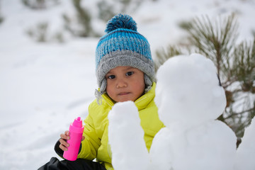 A toddler wearing winter clothes and beanie with paint bottle in hand, about to decorate a snowman.