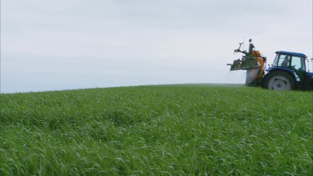 Medium Low Angle Still Shot Of A Blue Tractor Spraying Pesticide On Countryside Grass Field Against Grey Sky, UK