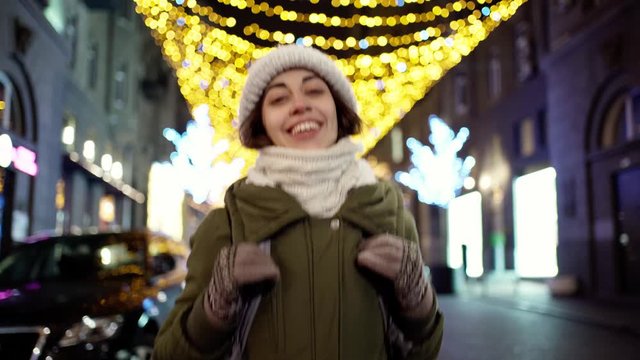 happy laughing young woman wearing warm winter clothes fast playfully walking night city street with beautiful bright Christmas illumination on many garlands background