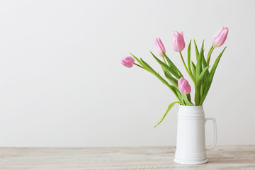 pink tulips in white ceramic jug on wooden table on background white wall