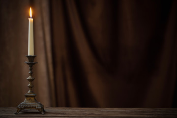 antique candlestick with burning candle on old wooden table on background brown velvet curtain