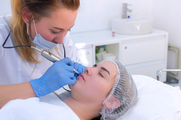 Cosmetologist making lips microblading procedure for girl using tattoo machine. Beautician is...