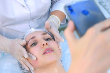 Obraz na płótnie Canvas Woman blogger was making facial massage shotting video on mobile phone talking with subscribers, closeup portrait. Cosmetologist massaging mature woman face in beauty clinic. Skincare procedure.